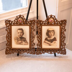May include: Two antique bronze-colored picture frames with ornate scrollwork and small floral accents. Each frame holds a sepia-toned portrait of a child. The frames are displayed on small black easels.