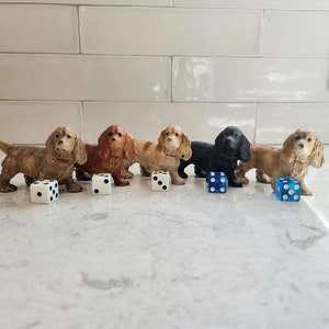 May include: A collection of six dog figurines in various colors, including brown, tan, and black, each with a red collar. Dice with white and blue dots are placed in front of each dog. The figurines are arranged on a light-colored surface.