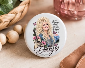 May include: A round white button featuring an illustration of Dolly Parton with blonde hair, a guitar, pink flowers, and butterflies. The text reads: If you see someone without a smile, give them one of yours. Dolly 1946-2026.