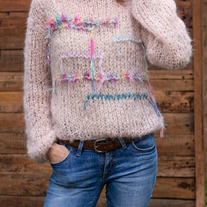 May include: A light pink, hand-knitted sweater with a crew neck and long sleeves. The sweater features colorful, textured embellishments across the chest and sleeves. The person is wearing blue jeans and a brown belt.