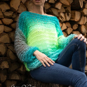 May include: A hand-knitted, ombre sweater in shades of teal, green, and yellow. The sweater has a loose, open-knit design and is made with a soft, fuzzy yarn. The sweater is worn by a person sitting on a tree stump.