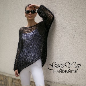 May include: A black, loose-fitting, open knit jumper with a scoop neck. The jumper is worn over white trousers with a patch on each knee. The model is wearing sunglasses and is standing against a white wall with stone accents. The text "Gerry Yap HANDKNITS" is visible in the lower right corner of the image.