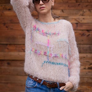 May include: A light pink, oversized sweater with colorful embroidered accents. The sweater has long sleeves and a crew neck with a light blue trim. The person in the image is wearing blue jeans and sunglasses.