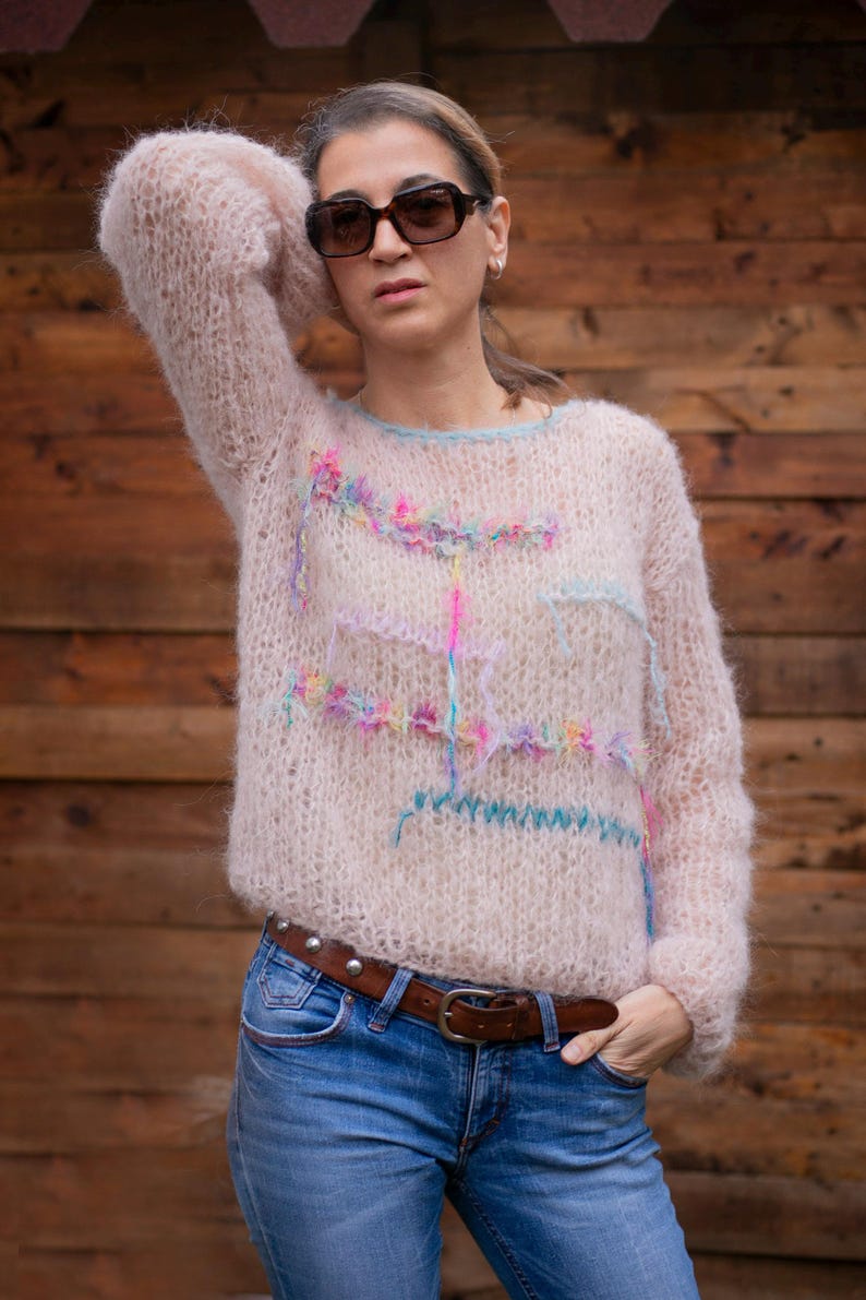 May include: A light pink, oversized sweater with colorful embroidered accents. The sweater has long sleeves and a crew neck with a light blue trim. The person in the image is wearing blue jeans and sunglasses.