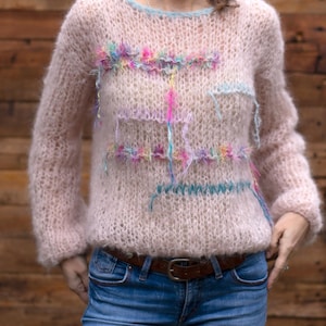 May include: A light pink, fuzzy sweater with a crew neck and long sleeves. The sweater features colorful embroidered accents in shades of pink, blue, and yellow. The model is wearing blue jeans and a brown belt.