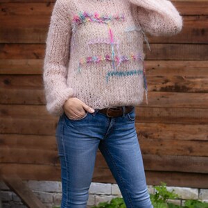 May include: A light pink, long-sleeved sweater with a crew neck and decorative colorful yarn accents. The sweater is paired with blue jeans and a brown belt. The model is wearing sunglasses.