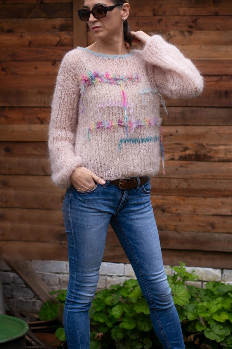 May include: A light pink, long-sleeved sweater with a crew neck and decorative colorful yarn accents. The sweater is paired with blue jeans and a brown belt. The model is wearing sunglasses.