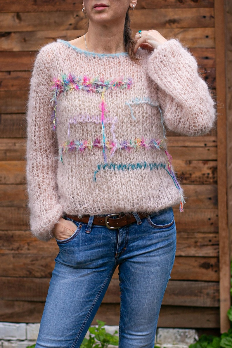 May include: A light pink, hand-knitted sweater with a crew neck and long sleeves. The sweater features colorful, textured embellishments across the chest and sleeves. The person is wearing blue jeans and a brown belt.
