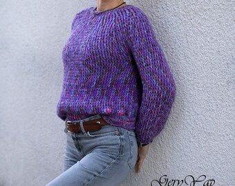 May include: A purple hand-knitted sweater with a round neck and long sleeves. The sweater has a textured knit pattern and is worn with blue jeans and a brown belt. A dark gray cap is worn on the head. The text "Gery Yap HANDKNITS" is visible.