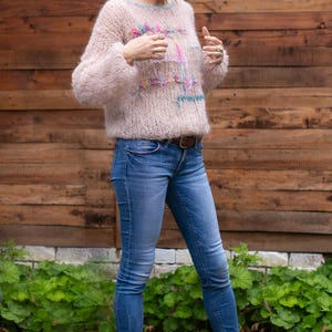 May include: A light pink, fluffy sweater with colorful embroidery is paired with blue jeans and gray clogs. The sweater has a crew neck and long sleeves. The jeans are fitted, and the clogs have a strap across the top.
