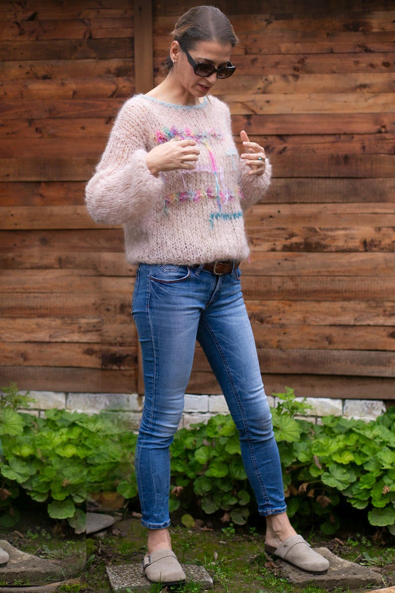 May include: A light pink, fluffy sweater with colorful embroidery is paired with blue jeans and gray clogs. The sweater has a crew neck and long sleeves. The jeans are fitted, and the clogs have a strap across the top.