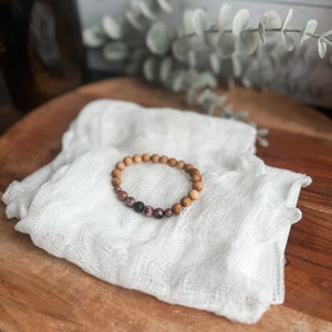 May include: A wooden bead bracelet with pink and black accent beads rests on a white cloth. The bracelet is displayed on a wooden surface, with a blurred background featuring a dark bottle and greenery.