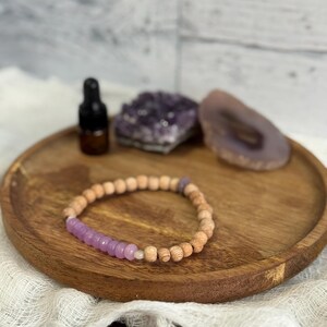 May include: A wooden beaded bracelet with a section of lavender-colored beads rests on a wooden tray. A small brown bottle, a cluster of amethyst crystals, and a slice of agate are also on the tray. The tray sits on a white cloth.