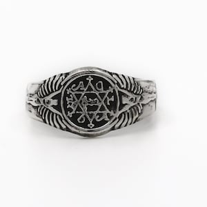 May include: A silver ring with a black enamel center featuring a six-pointed star with a symbol inside. The ring has two wings on either side of the star.