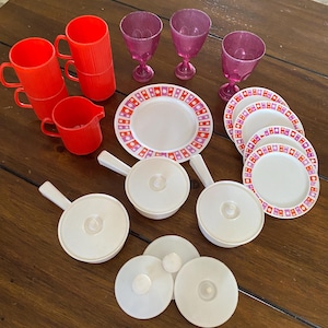 May include: A set of red and white plastic toy dishes and cups, including a pitcher, bowls, plates, and wine glasses. The plates have a pink and white geometric pattern.