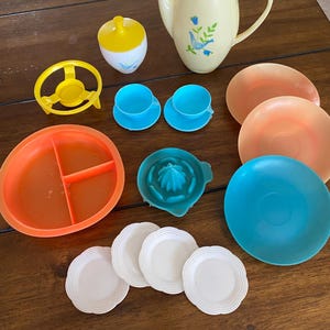 May include: A vintage plastic toy tea set with a yellow teapot, sugar bowl, and creamer. The set also includes a yellow stand, a blue and white pitcher, two blue cups and saucers, three white plates, a blue bowl, an orange bowl with three sections, and a green citrus squeezer.