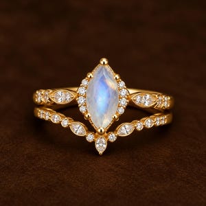 May include: A gold ring set featuring a central marquise-cut moonstone surrounded by a halo of small round white stones. The band is accented with marquise and round stones, and it is paired with a matching V-shaped chevron band.
