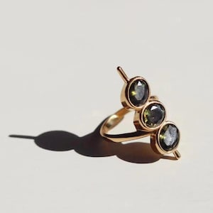 May include: A gold-colored ring featuring three dark green gemstones. The ring has a unique design with the gemstones set in individual bezels. The ring is set against a white background, with a shadow cast to the left.