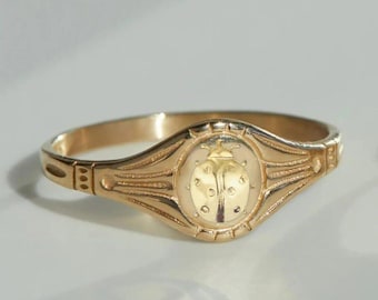 Può includere: Un anello dorato con un design dettagliato di coccinella al centro. L'anello ha una forma ovale con dettagli decorativi lungo la fascia. La coccinella è raffigurata con piccoli punti e un corpo strutturato.