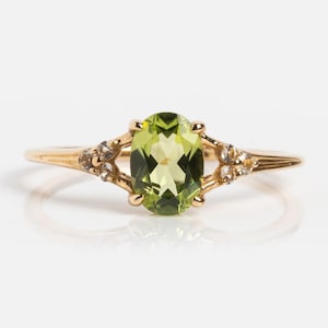 May include: A gold ring featuring a large, oval-cut peridot gemstone in a light green hue. The ring has small accent stones on either side of the peridot and a delicate band. The ring is set against a white background.