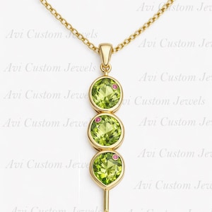 May include: A gold necklace featuring a pendant with three round, light green gemstones, each accented with a small pink gem. The pendant is suspended from a delicate gold chain. The text "Avi Custom Jewels" is visible in the background.
