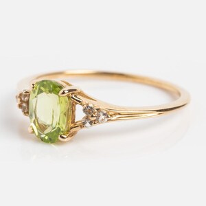 May include: A gold ring featuring a light green oval gemstone, flanked by smaller accent stones. The ring has a delicate band and a classic, elegant design. The gemstone is a vibrant, eye-catching color.