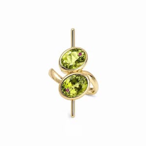 May include: A gold ring featuring two oval-cut lime green peridot gemstones set vertically. Each stone is accented with a tiny, round, deep pink gemstone. The metal band has a polished finish and extends slightly beyond the top and bottom stones.