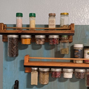 May include: Two wooden spice racks with metal rails, mounted on a blue wall. The top rack has six glass jars filled with spices and three bottles with green lids. The bottom rack has eight glass jars filled with spices. The jars are hanging from the rails. The top rack has a label that reads "Mushroom & Company Umami Multipurpose Blend".
