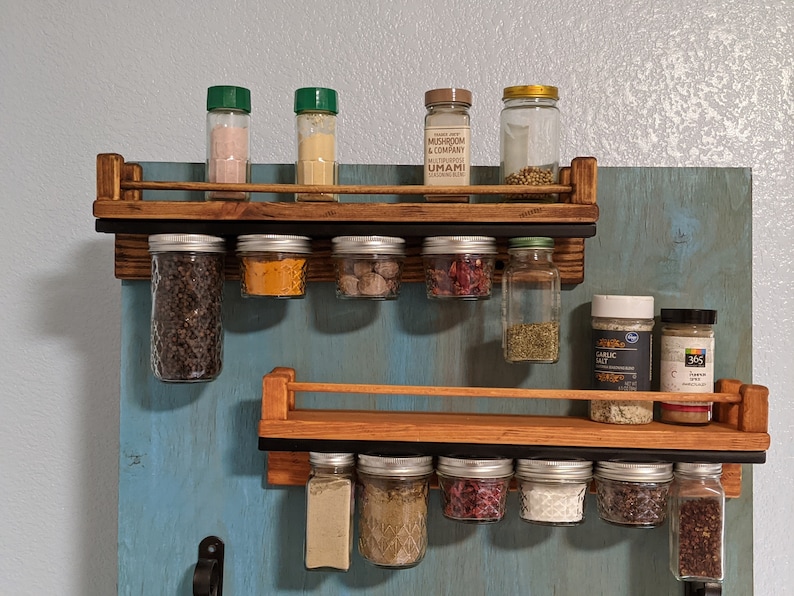 May include: Two wooden spice racks with metal rails, mounted on a blue wall. The top rack has six glass jars filled with spices and three bottles with green lids. The bottom rack has eight glass jars filled with spices. The jars are hanging from the rails. The top rack has a label that reads "Mushroom & Company Umami Multipurpose Blend".