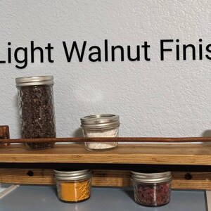 May include: A light walnut finish spice rack with four glass jars filled with spices. The rack has a black metal bar and is mounted to a wall.