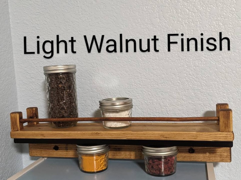 May include: A light walnut finish spice rack with four glass jars filled with spices. The rack has a black metal bar and is mounted to a wall.
