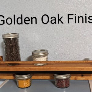 May include: A wooden spice rack with a black metal bar and four glass jars filled with spices. The rack is labeled "Golden Oak Finish".