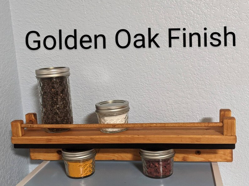 May include: A wooden spice rack with a black metal bar and four glass jars filled with spices. The rack is labeled "Golden Oak Finish".