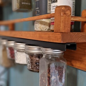 May include: A wooden spice rack with a black metal bar for hanging jars. The rack is mounted on a wall and has several glass jars filled with spices hanging from the bar. The jars are labeled with different spices, including "Pumpkin Spice".