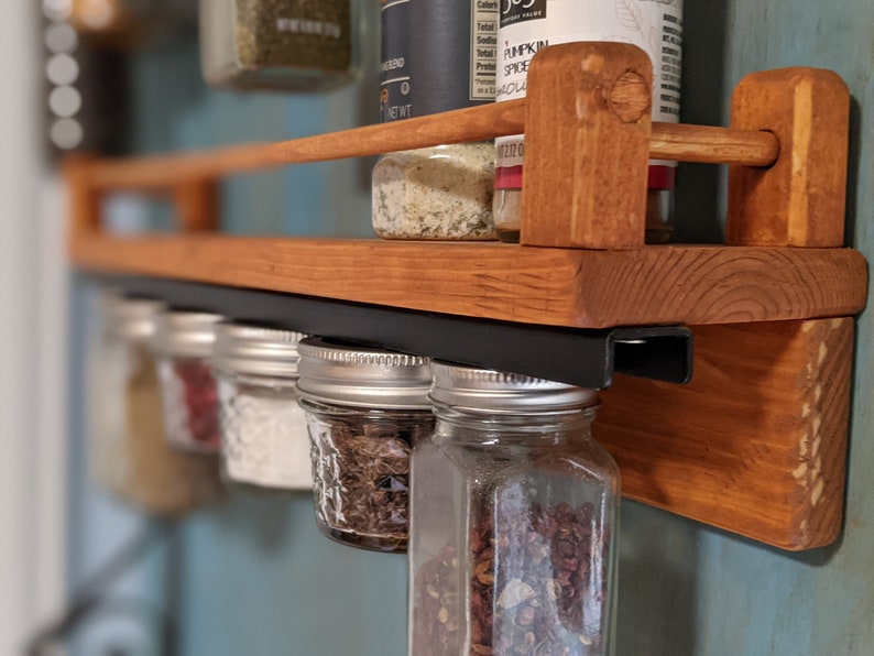 May include: A wooden spice rack with a black metal bar for hanging jars. The rack is mounted on a wall and has several glass jars filled with spices hanging from the bar. The jars are labeled with different spices, including "Pumpkin Spice".