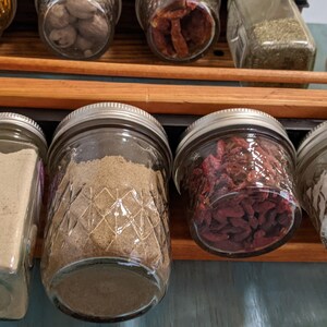 May include: A close-up of a spice rack with various spices in glass jars. The jars are filled with different spices, including ground spices, dried fruit, and herbs. The jars are arranged in a row on a wooden shelf.