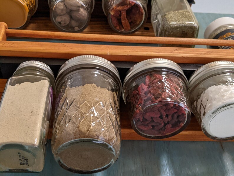 May include: A close-up of a spice rack with various spices in glass jars. The jars are filled with different spices, including ground spices, dried fruit, and herbs. The jars are arranged in a row on a wooden shelf.