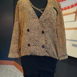 May include: A brown and black patterned blouse with a double-breasted button closure. The blouse is layered over a black turtleneck and black pants.