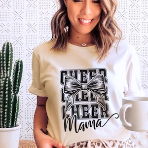 Puede incluir: Una camiseta blanca con un gráfico negro que dice "Cheer Cheer Cheer Mama" con un gran lazo negro en el centro.