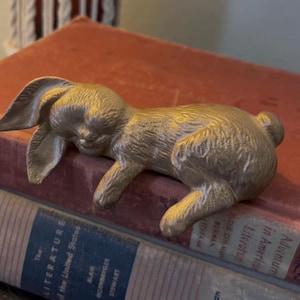 Puede incluir: Una figura de metal en tono dorado de un conejo durmiendo con detalles de pelaje texturizado, orejas largas y una cola pequeña, en posición horizontal. El conejo tiene una superficie lisa y reflectante, fundida en una sola pieza.