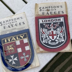 Può includere: Due stemmi Sampson's Souvenir Badges in confezioni di plastica trasparente. Uno stemma riporta la parola ITALY sopra uno scudo con quattro quadranti contenenti un leone e delle croci. L'altro stemma riporta la parola LONDON sopra uno stemma.