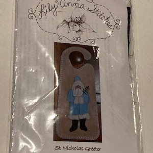 Puede incluir: Un paquete de patrón de manualidades de Lily Anna Stitches titulado St Nicholas Greeter Pattern LAS942. El empaque presenta una imagen impresa de un colgador de puerta de fieltro beige en forma de gancho con una figura de San Nicolás en abrigo azul.