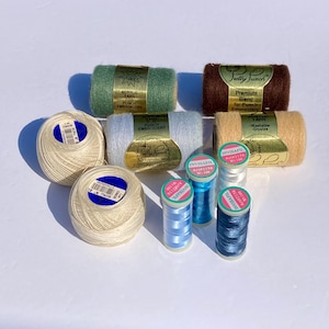 May include: A collection of craft supplies including two cream-colored yarn balls, three rectangular spools of acrylic yarn in green, brown, and light blue, and three small spools of Invisafil thread in light blue, medium blue, and white.