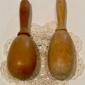 Puede incluir: Dos maracas de madera con cuerpos redondeados y lisos y mangos delgados. La maraca de la izquierda presenta un acabado marrón medio cálido, mientras que la de la derecha muestra una veta de madera natural más clara, desgastada y texturizada.