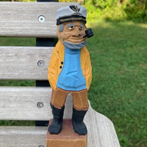 May include: A hand-carved wooden figurine of a sailor wearing a white captain's hat, a yellow jacket, a light blue shirt, brown trousers, and black boots. The figure has a grey beard and is holding a small black pipe in its mouth, standing on a square wooden base.