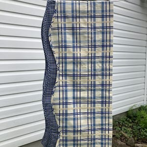 May include: A rectangular fabric runner featuring a cream-colored base with a blue and yellow plaid pattern on the front side. The reverse side displays a dark blue fabric with a small white grid pattern, visible along the wavy edge.