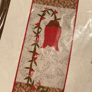 May include: A rectangular fabric wall hanging featuring a central light gray panel with a delicate white floral pattern. A large red bell-shaped flower on a green vine with smaller red blossoms curves along the left side. Brown patterned borders frame the top and bottom.