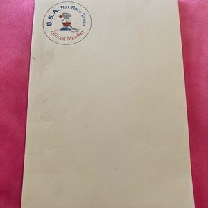 May include: A rectangular off-white notepad featuring a circular graphic in the top left corner. The graphic depicts a cartoon mouse wearing a red shirt and blue shorts, with the text U.S.A. Rat Race Team Official Member printed around it.