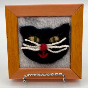May include: A needle-felted black cat face with yellow-green eyes, a bright red nose, and white yarn whiskers, set against a light gray felt background. The artwork is encased in a square, light-toned wooden frame with a silver-colored metal stand.