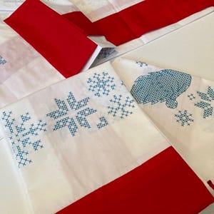 Op de afbeelding: Een witte stoffen tafelloper met een rode rand en blauwe kruissteek borduurpatronen van sneeuwvlokken en een kabouter. De tekst "Table Topper Motif" is op de stof gedrukt. Het ontwerp bevat geometrische vormen en een figuur met een baard.